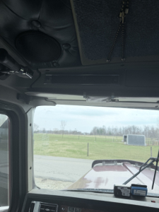 Kenworth W900L truck cab interior front view showing dashboard, windshield wipers, sun visor, and ceiling vent; exterior road and field visible through windshield.