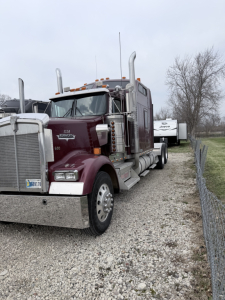 Kenworth W900L conventional sleeper truck in a used condition with chrome bumper, tall exhaust stacks, maroon cab, and trailers visible in background.