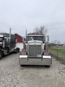 Front view of a 1999 Kenworth W900L conventional sleeper truck showing a large chrome grille, dual tall exhaust stacks, amber clearance lights, and a front bumper. License plate visible on bumper.