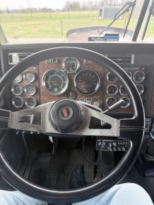 Steering wheel and gauge cluster in a Kenworth W900L interior showing multiple round gauges and a woodgrain dash