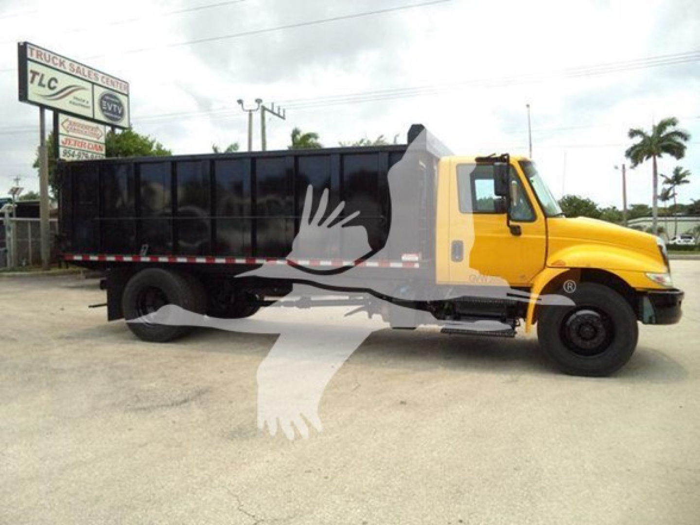 Truck showing a yellow cab and black dump bed with a large watermark graphic in front. Side text on truck not legible; background shows dealership lot with TLC sign.