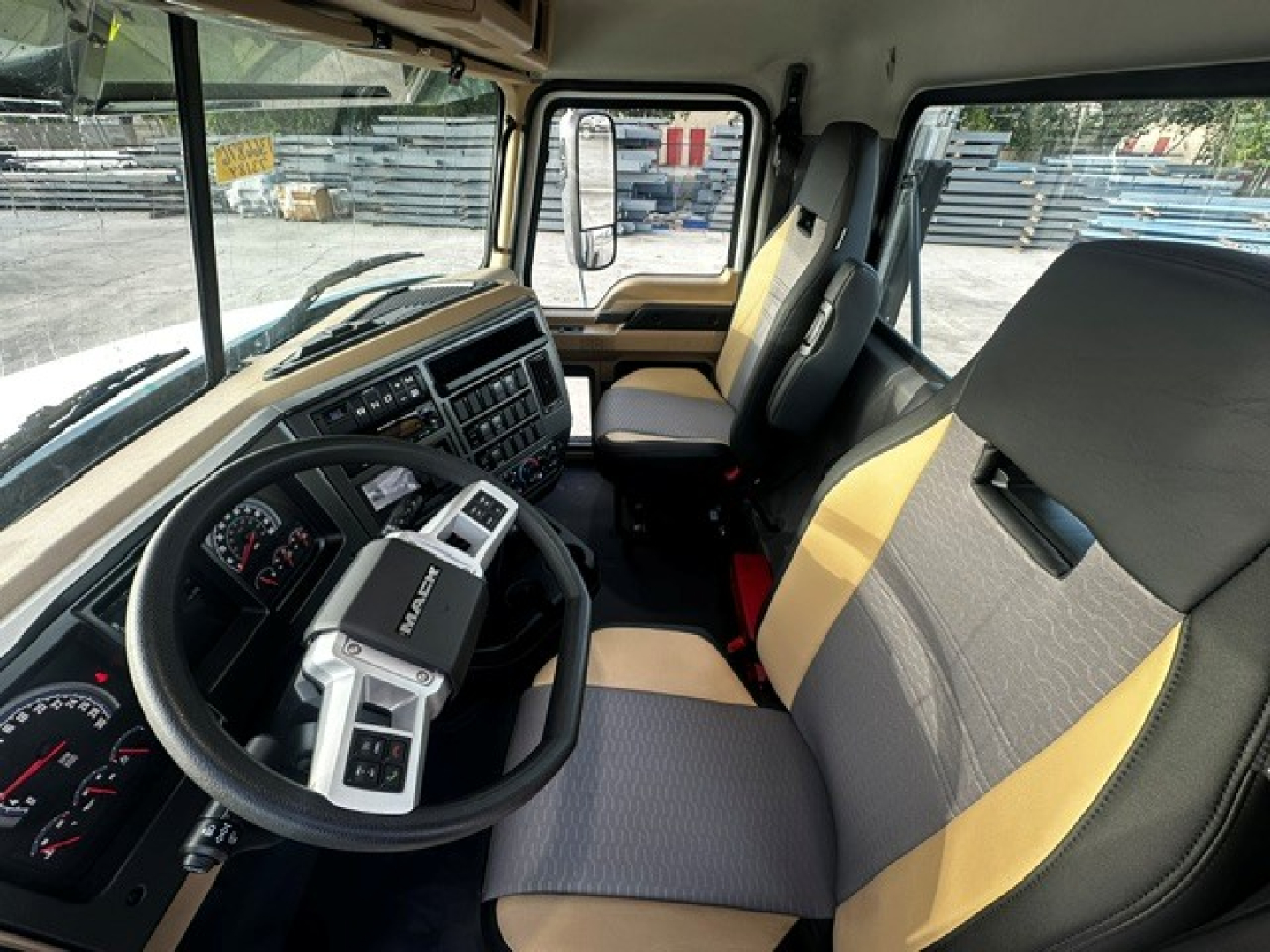 Truck cab interior showing steering wheel, instrument cluster, and driver and passenger seats. Dashboard with controls and gear selector, Mack logo visible on steering wheel.