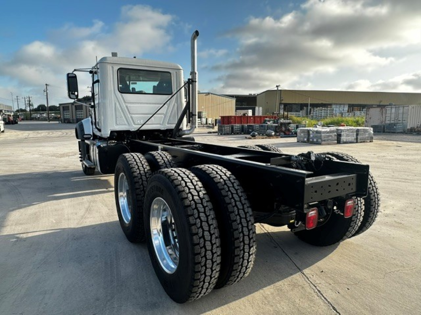 Truck chassis with dual rear axles, cab in front, Mack GR64F configuration. View from rear-left showing frame, dual rear tires, exhaust stack, and fifth-wheel area.