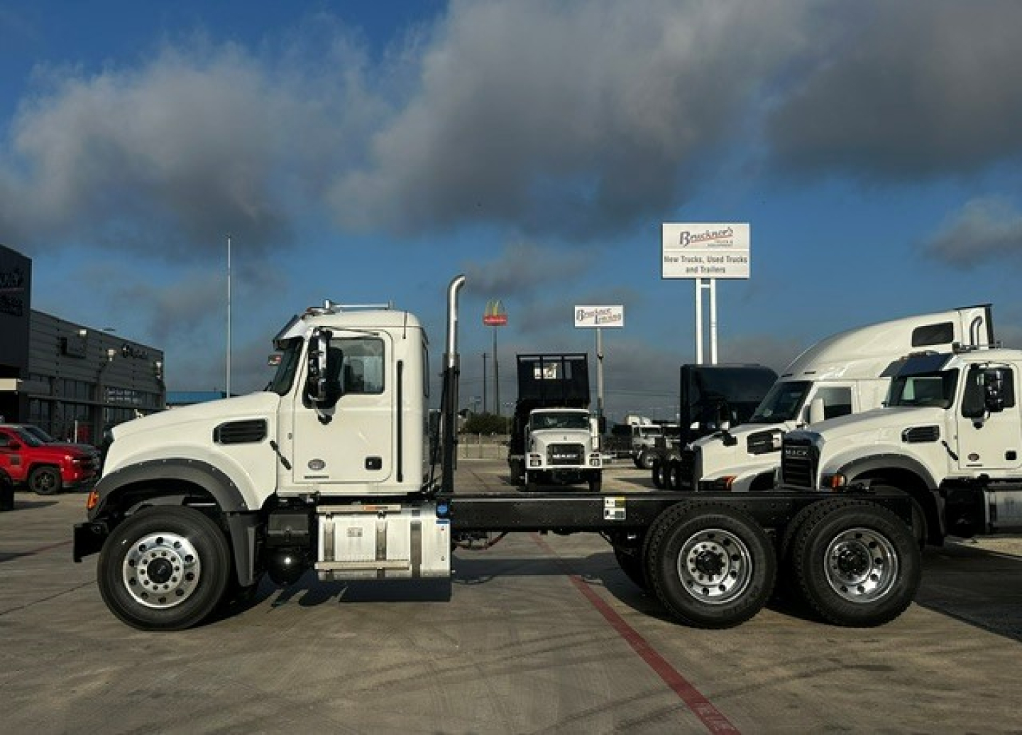 White Mack GR64F truck cab with chassis and rear axles visible, side view in a dealership lot with other white trucks in background.
