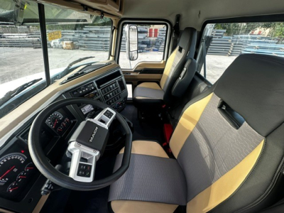 Truck cab interior showing steering wheel, instrument cluster, and driver and passenger seats. Dashboard with controls and gear selector, Mack logo visible on steering wheel.