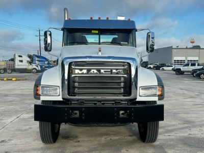 Front view of a white Mack truck with large grille reading MACK, bumper guard, dual side mirrors; vehicle in a lot with other trucks.