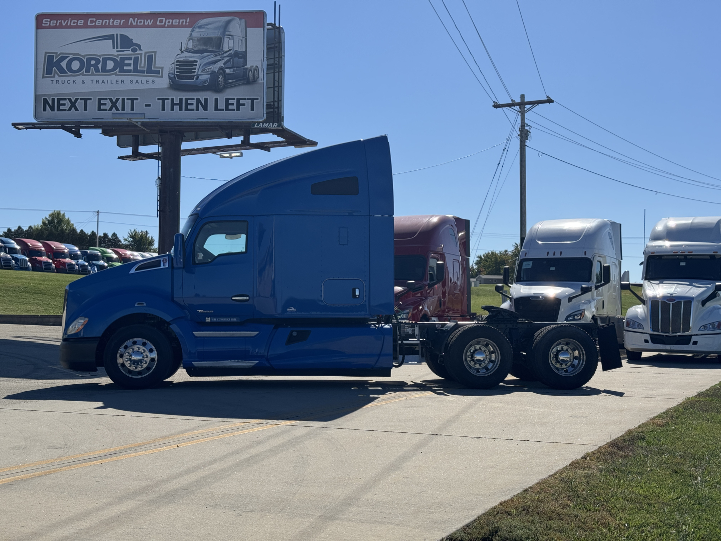 2015 Kenworth T680 tractor-trailer unit in blue with sleeper cab, attached to a black trailer. Billboard and other trucks in background.