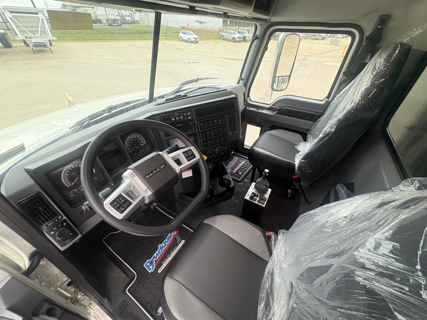 Truck cab interior of a 2027 Mack GR84F showing steering wheel, dashboard with gauges, center console, and driver seat covered in plastic.