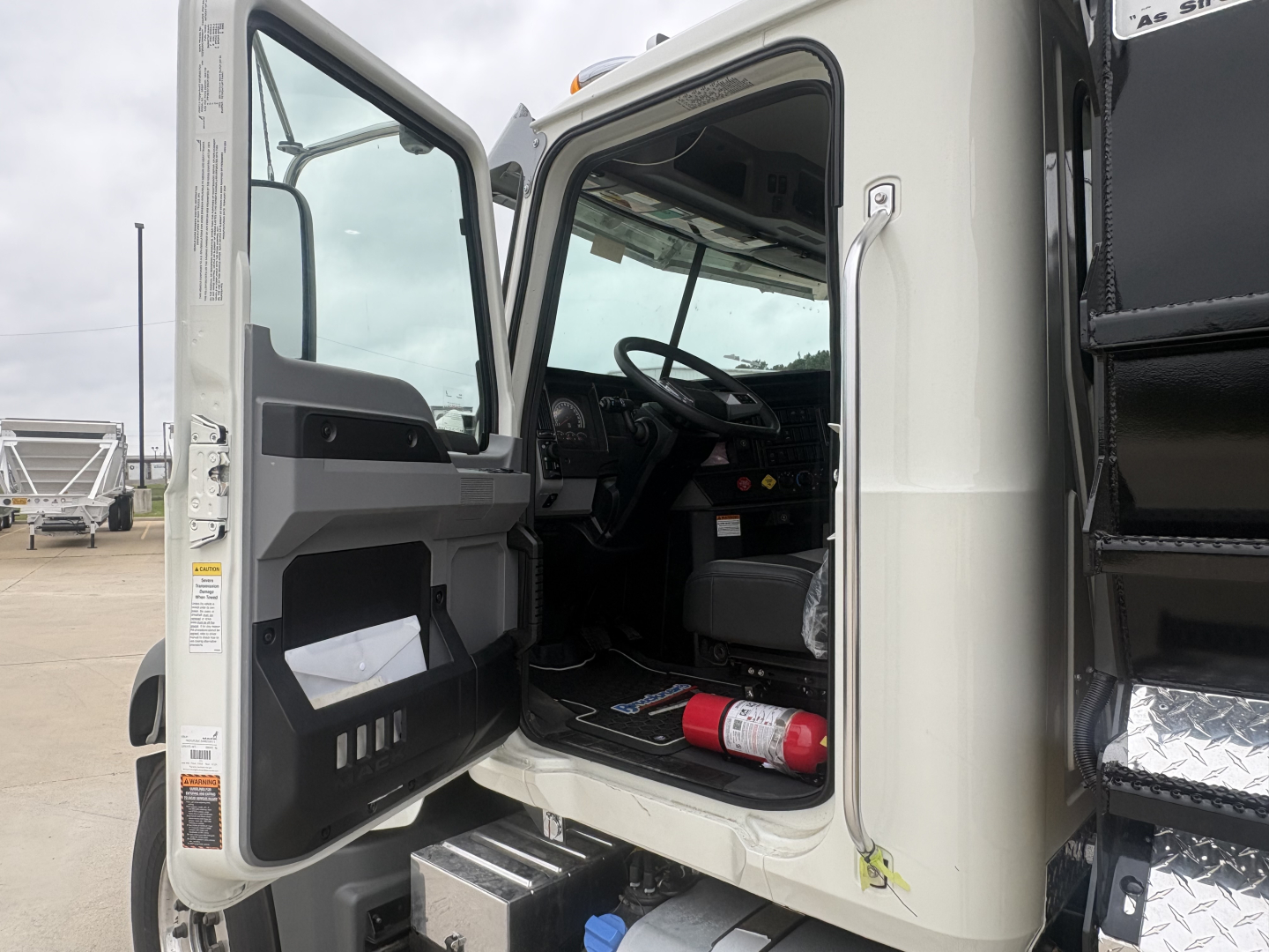 Cabin of a new Mack GR84F dump truck with door open, showing interior dashboard, steering wheel, and a red fire extinguisher under the driver seat.