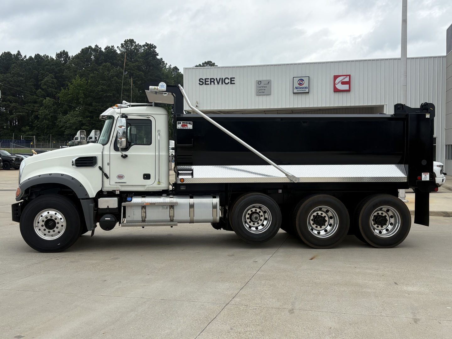 Dump truck side view showing white cab and black dump body, Mack GR84F style with silver fuel tanks and triple rear axles in front of service building.