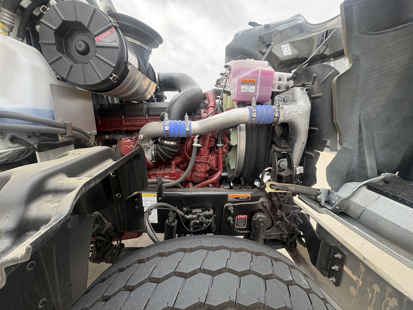 Mack GR84F engine bay showing red engine block, blue hose clamps, pink coolant reservoir, and large front tire in view.