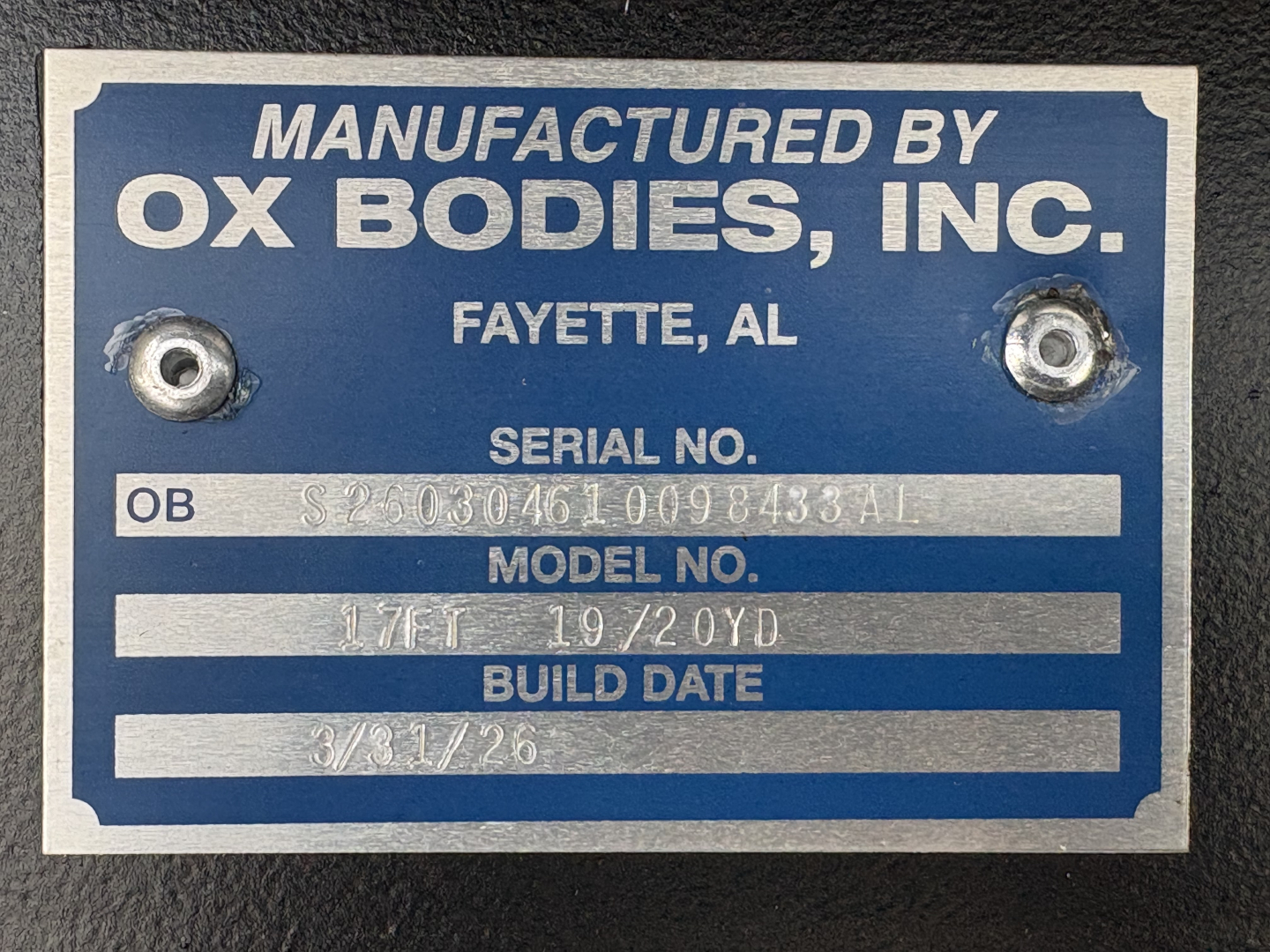 Manufacturer plate showing model and build data: ‘Manufactured by OX Bodies, Inc.’, Fayette, AL with serial number, model no, build date 3/31/26; two screw holes.