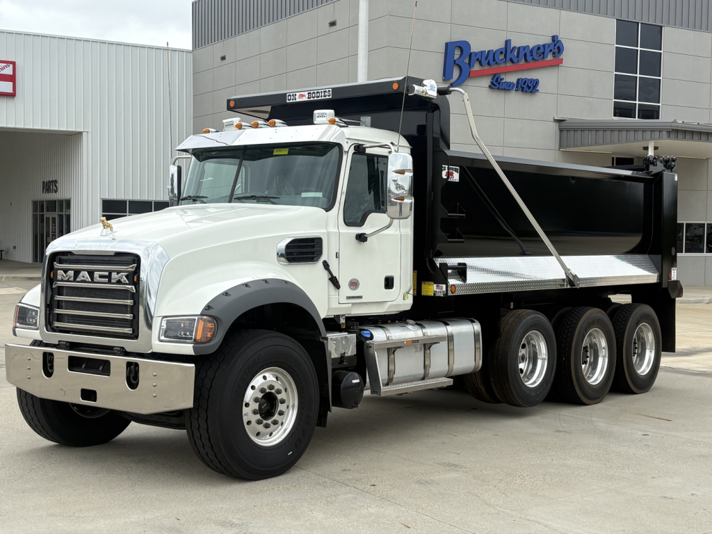 Dump truck showing Mack GR84F with white cab and black dump bed, side view, three-axle configuration, Brackner's dealership in background.