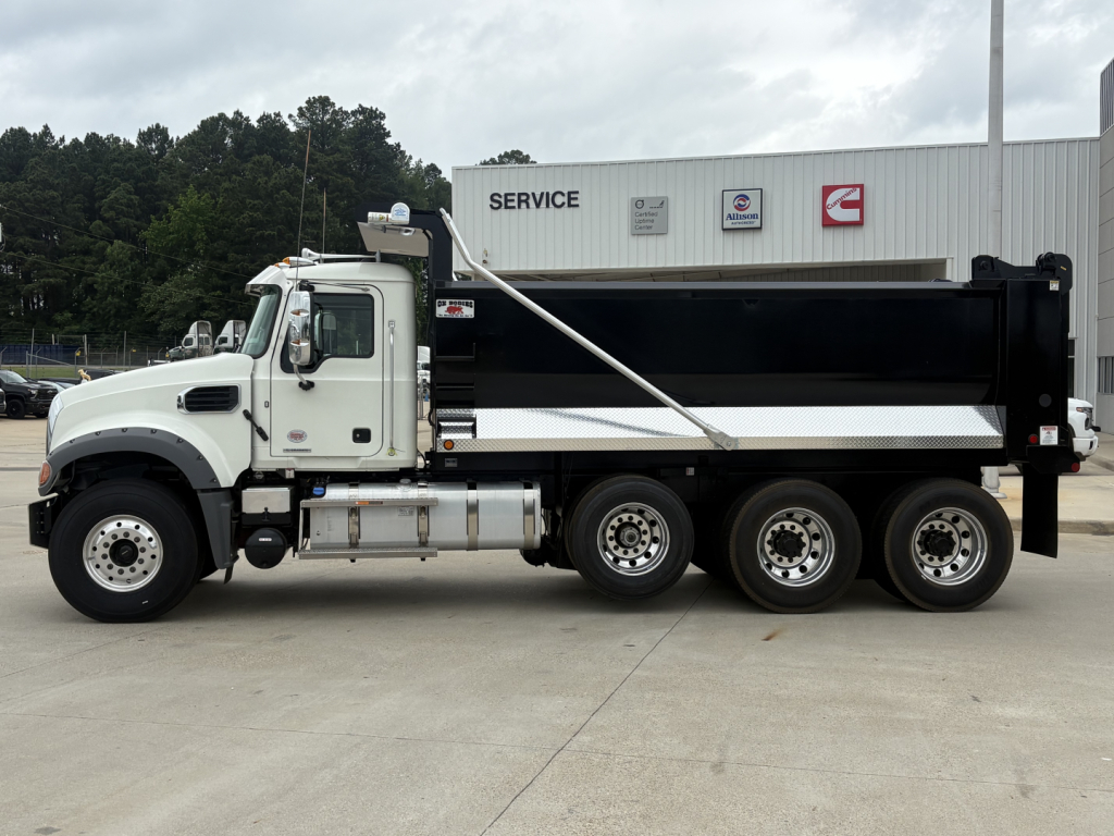 Dump truck side view showing white cab and black dump body, Mack GR84F style with silver fuel tanks and triple rear axles in front of service building.