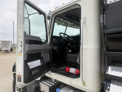 Cabin of a new Mack GR84F dump truck with door open, showing interior dashboard, steering wheel, and a red fire extinguisher under the driver seat.