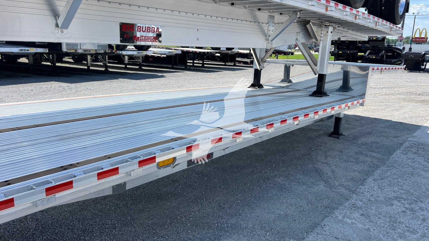 Flatbed trailer side view showing dropped deck and aluminum surface with reflective tape and support legs.