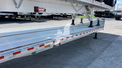 Flatbed trailer side view showing dropped deck and aluminum surface with reflective tape and support legs.