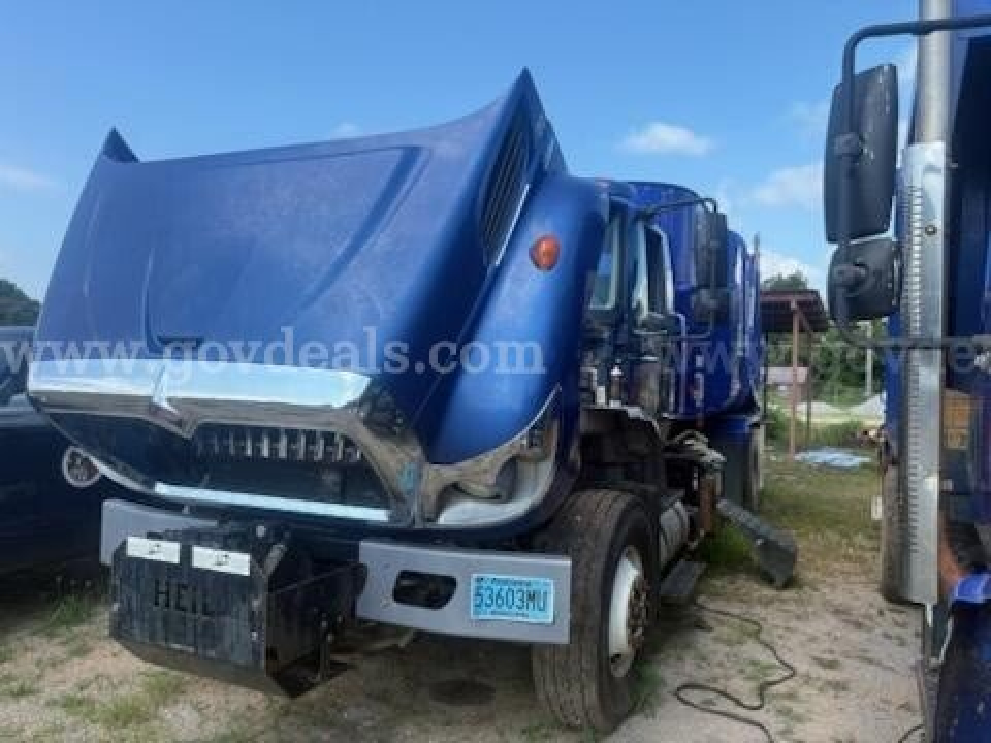 Truck cab with blue hood raised showing front bumper and side mirror; license plate area and blue state tag; visible text includes 12-digit license-like tags and website watermark; no clear engine text visible.