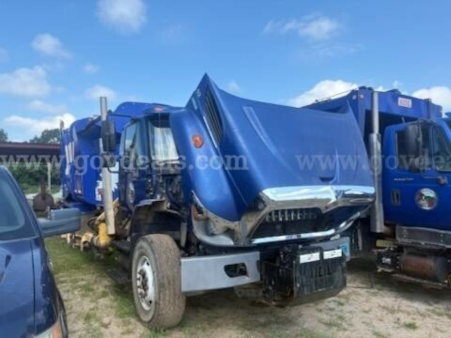 International 7500 garbage truck cab and front end with blue body and front bumper; visible damaged hood and grille, side mirrors, and large tires.