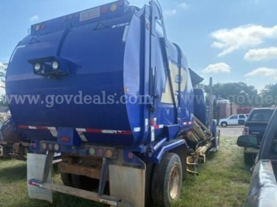 Garbage truck rear and side view with blue body and recycling compactor, dual wheels, visible warning lights and reflective tape.