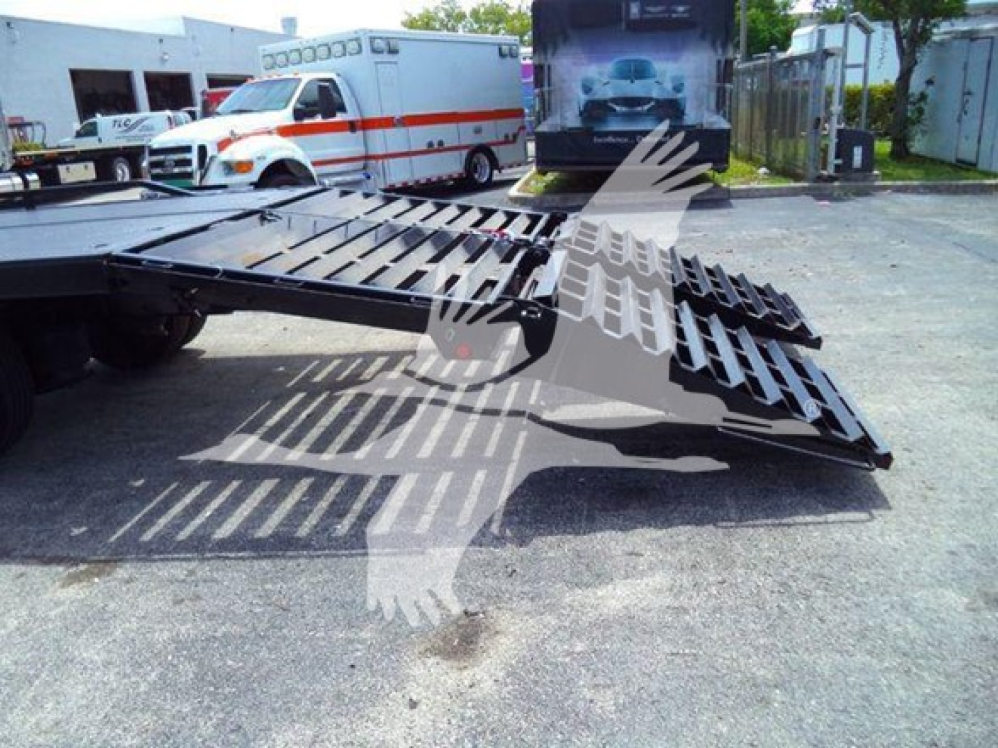 Flatbed truck with a folded gooseneck ramp on a Freightliner chassis, outdoor yard with ambulances in background.