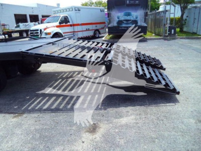 Flatbed truck with a folded gooseneck ramp on a Freightliner chassis, outdoor yard with ambulances in background.