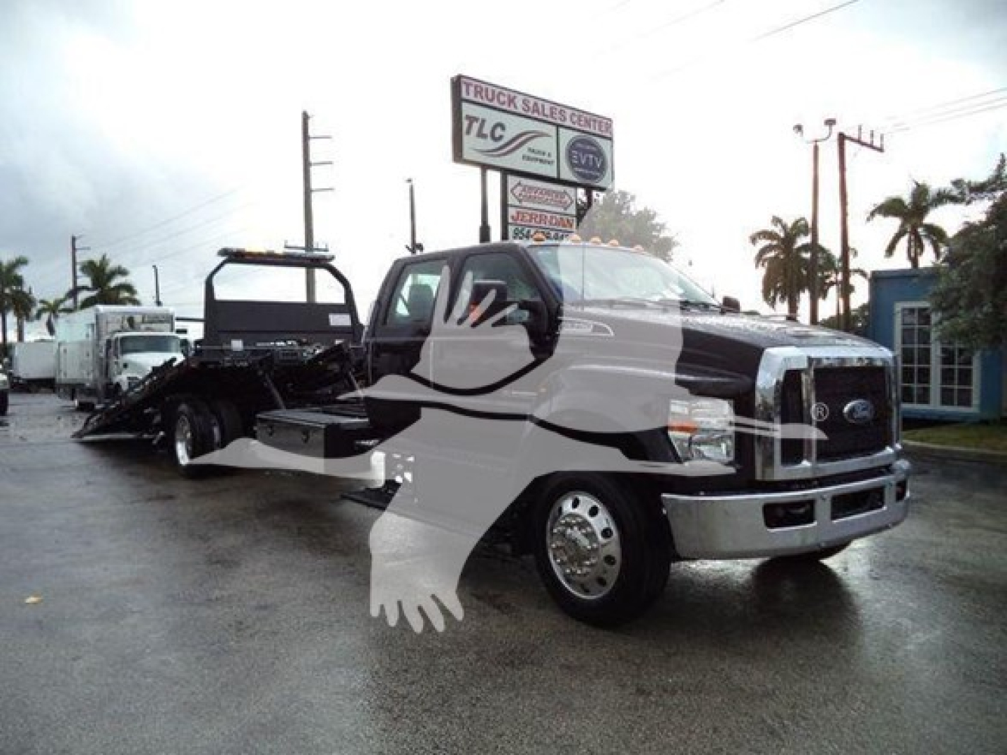 Ford F-750 rollback tow truck in view showing cab and flatbed with towing equipment; front grille and headlights visible; dealership lot in background.