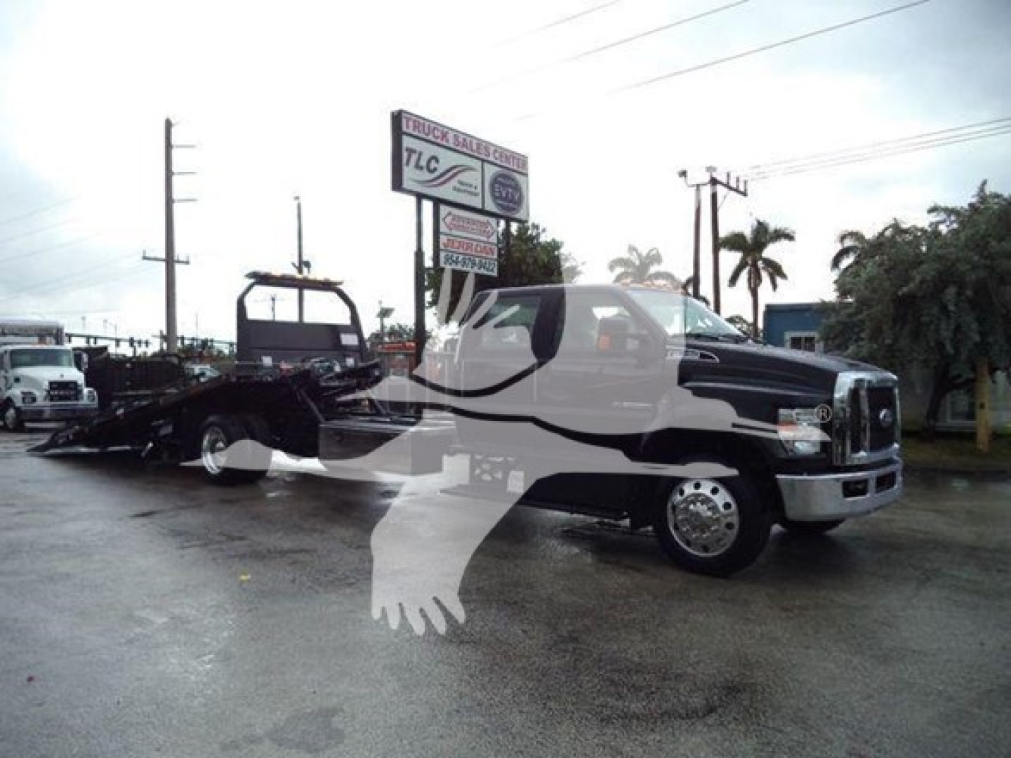2024 Ford F750 rollback truck with tow bed visible, side view showing cab and front grille, dealership sign in background.
