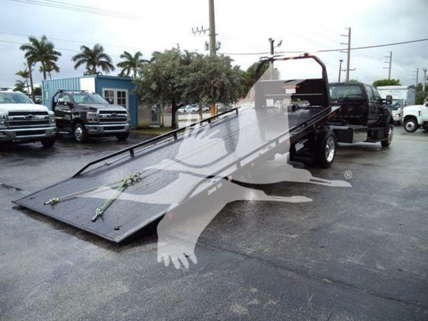 Ford F750 rollback truck with folded flatbed in foreground, tow truck bed and rear dual wheels visible, dealership lot in background.