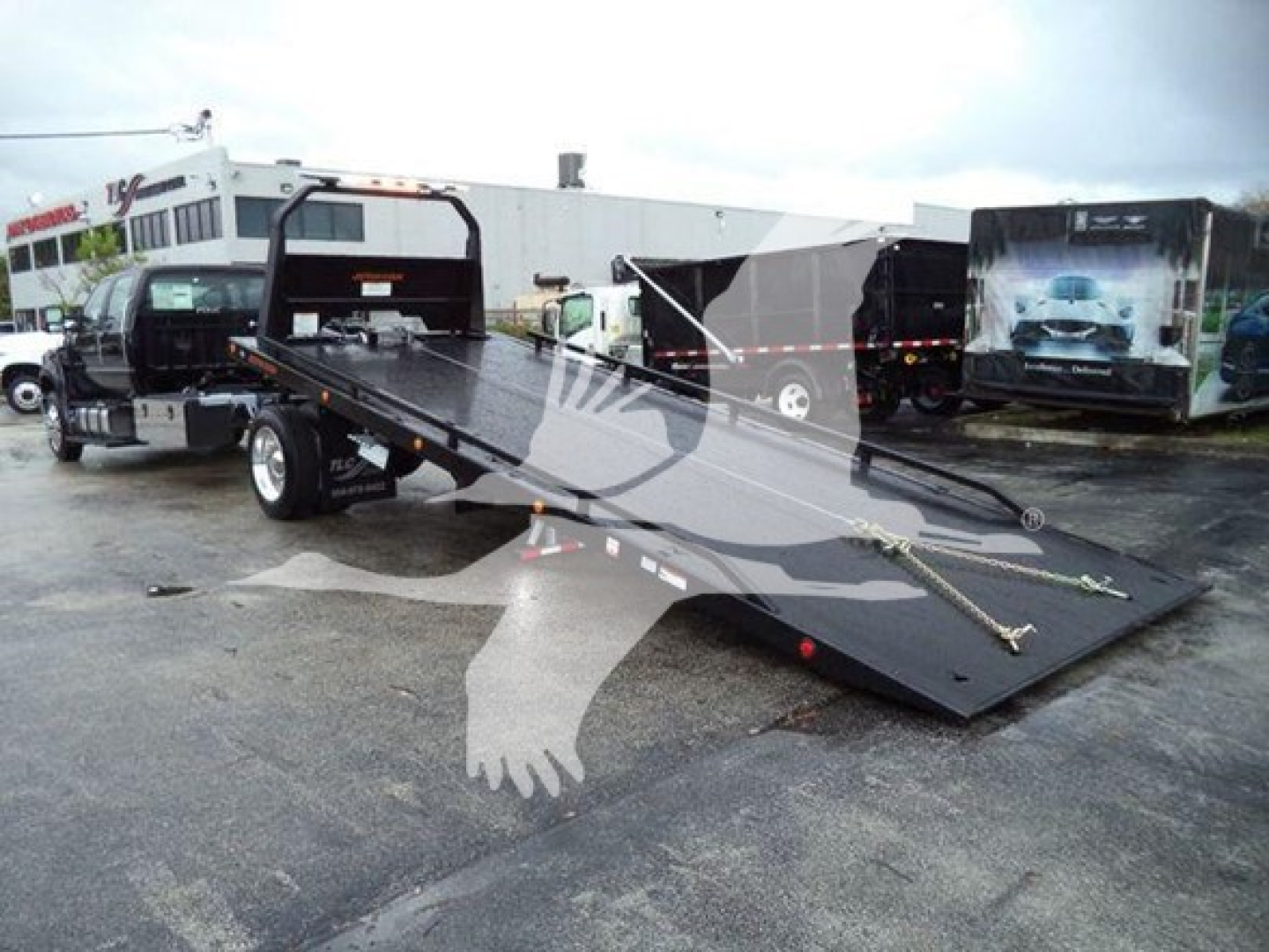 Flatbed rollback tow truck in parking lot with ramps extended and surrounding trailers.
