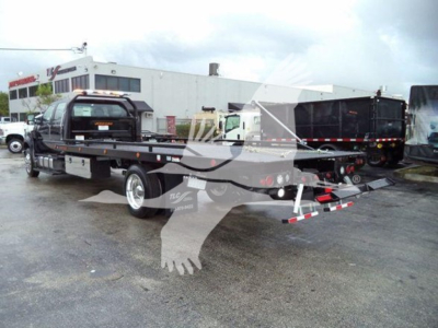 Flatbed rollback tow truck with a Ford F750 cab on a dealership lot; visible side text includes no clear legible labels; general equipment and vehicles in background.