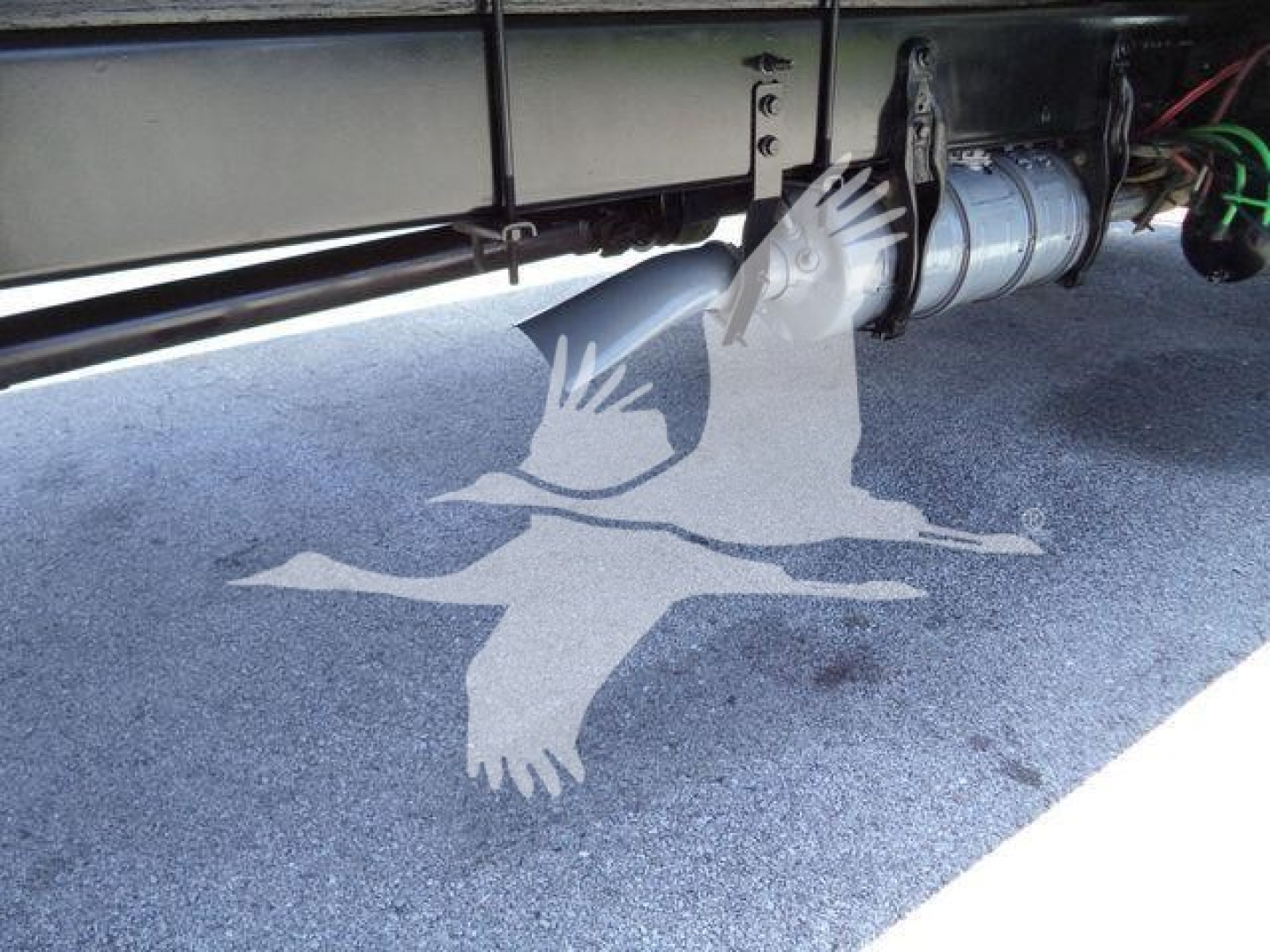 Truck undercarriage showing drivetrain and exhaust components mounted to frame; visible metal piping and a cylindrical exhaust/muffler assembly with wiring nearby.