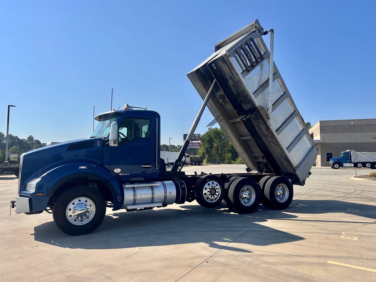 Blue Kenworth dump truck with raised dump bed, side view showing cab, front wheel, dual rear axles, and hydraulic dumping mechanism.