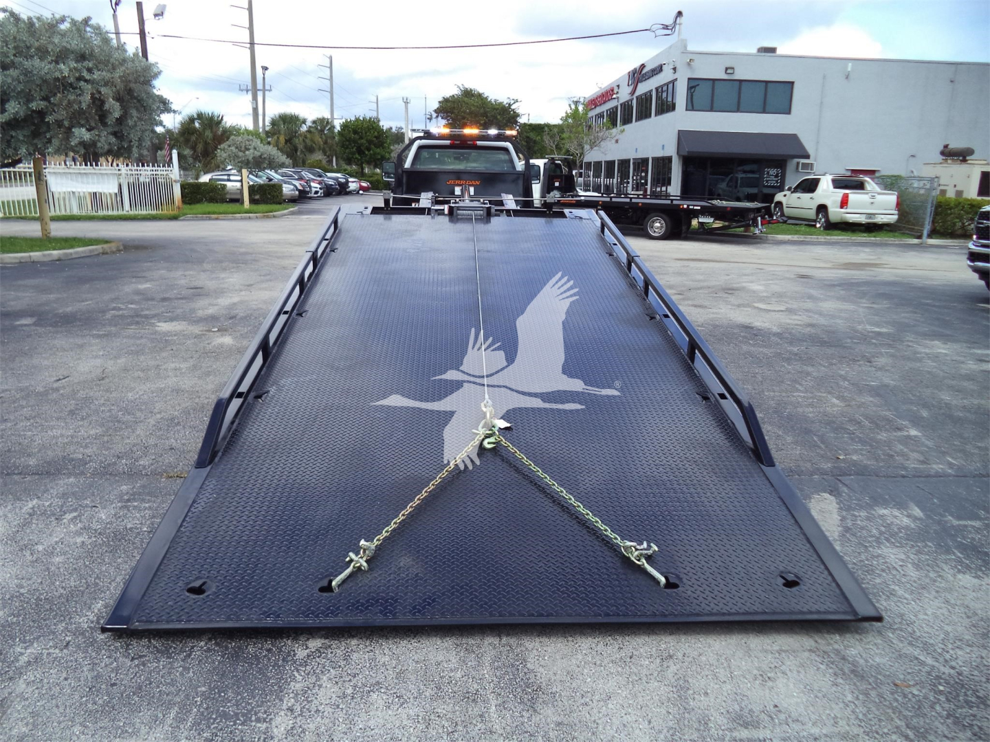 Truck bed of a rollback tow truck with a bird silhouette decal on the metal surface, safety chains across the bed, and surroundings including a building and parked vehicles.