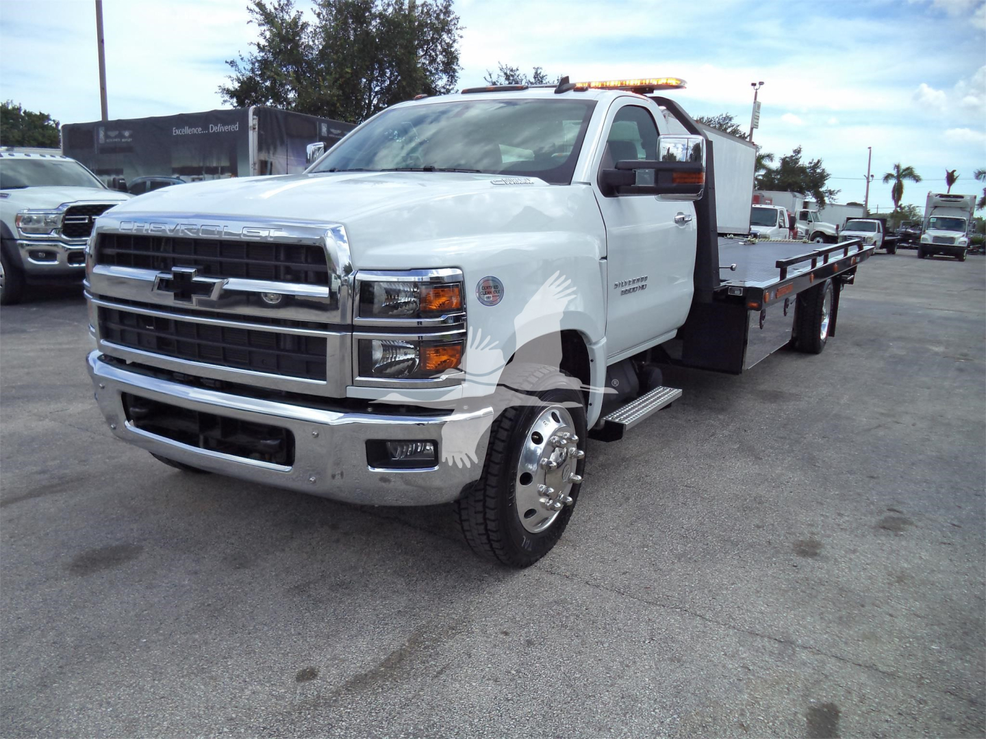 Chevrolet Silverado 6500HD rollback tow truck in white, front grille and chrome bumper, dual rear wheels, flatbed with wheel lift in background, dealership lot.