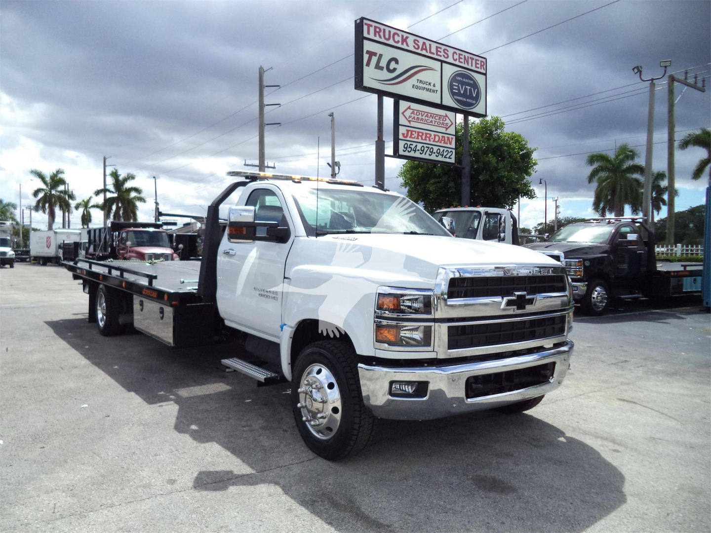 White Chevrolet Silverado 6500HD tow truck/rollback with long flatbed, front chrome bumper, diesel engine, in dealership lot with TLC Truck Sales Center sign.