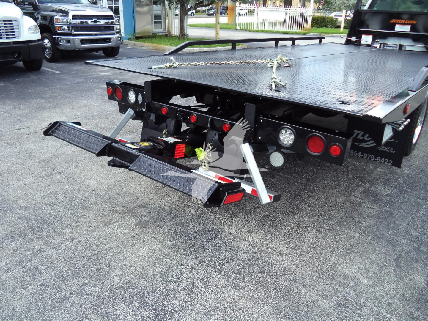 Rear view of a rollback tow truck bed with hydraulic wheel lift and fold-down ramps, black flatbed with rear lights and chain setup.