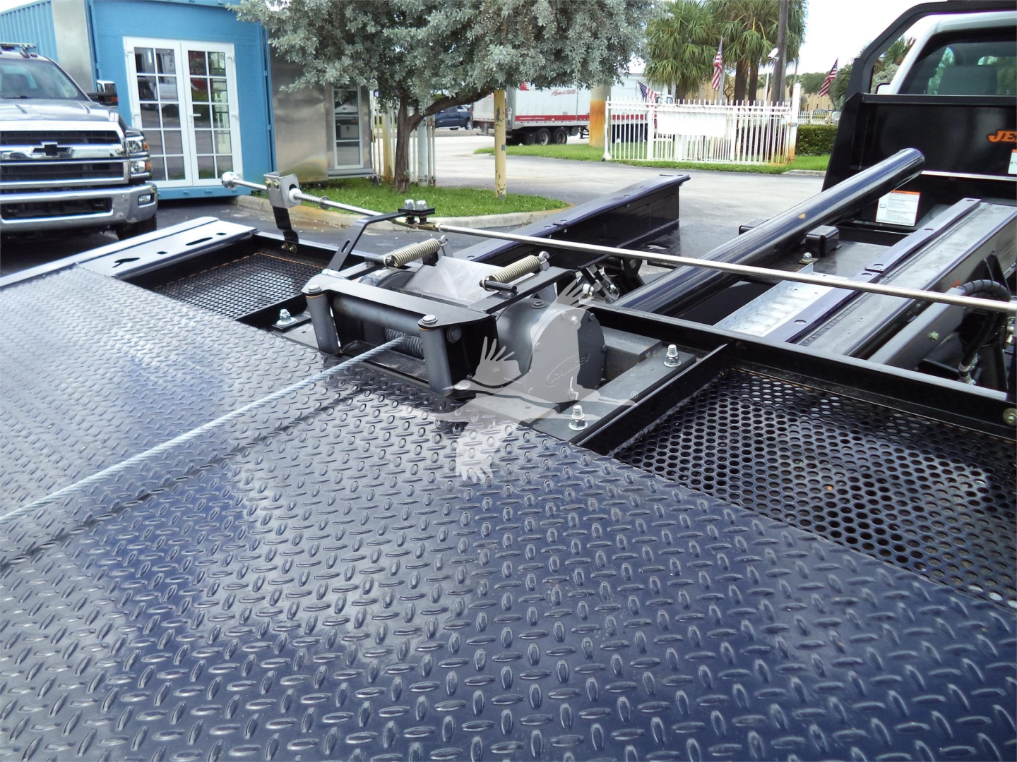 Truck bed with hydraulic wheel-lift mechanism and diamond plate surface; metallic components and rails visible on a rollback truck chassis.