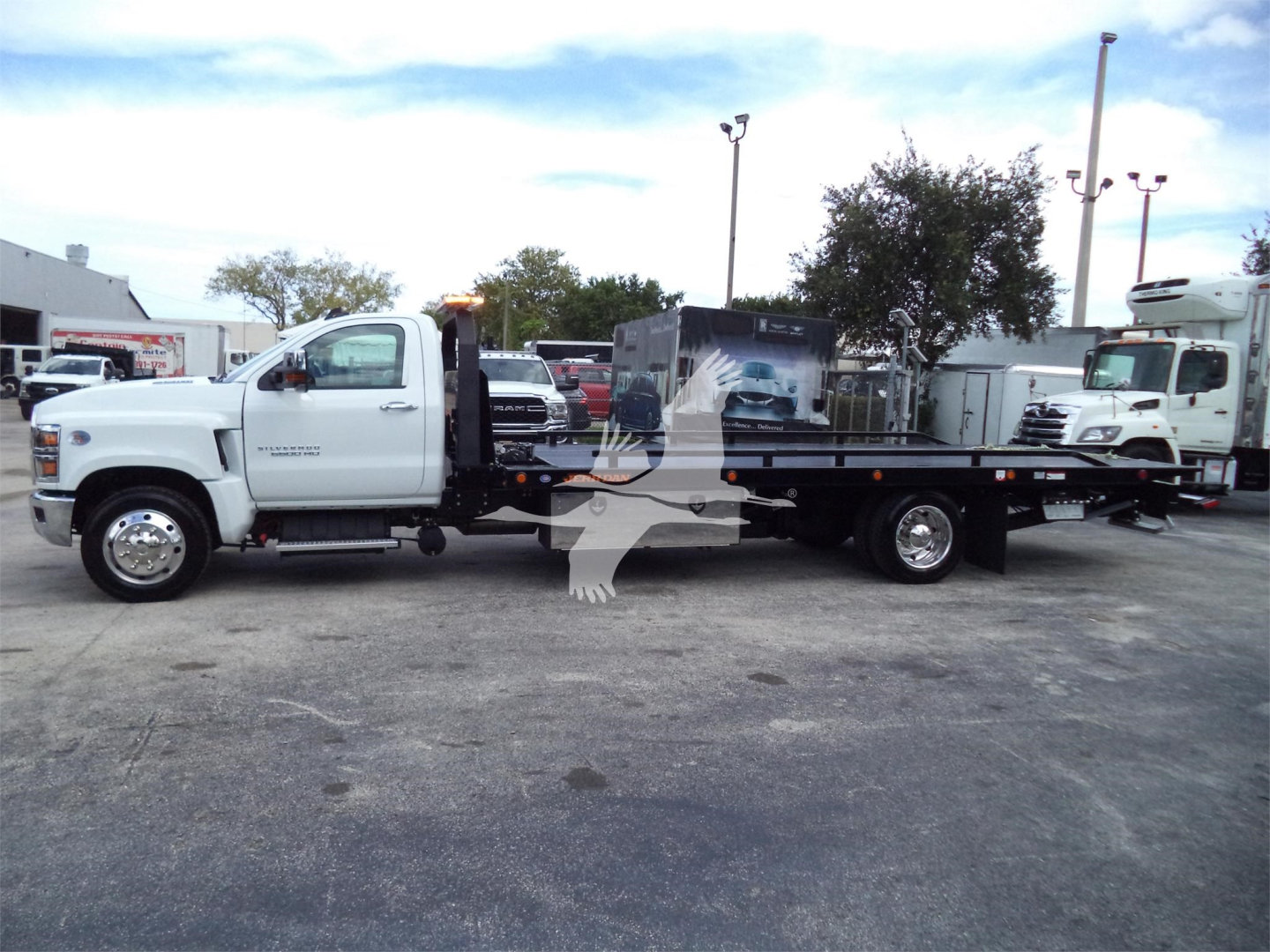 Truck side view of a 2023 Chevrolet Silverado 6500HD rollback tow truck showing flatbed with machinery in background.