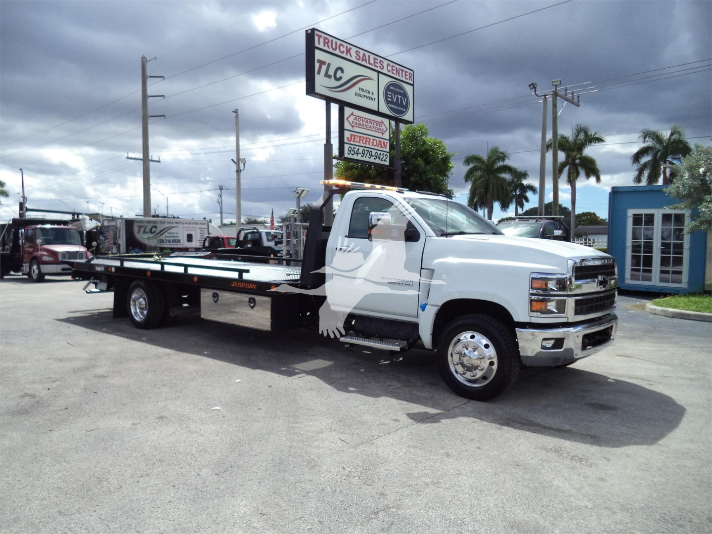 Truck with flatbed tow body in a lot; side shows no readable labels; background signage includes TLC Truck Sales Center and a blue building.