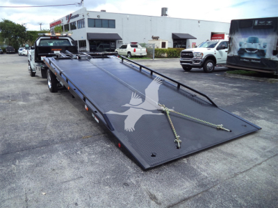 Truck bed rollback trailer with a long flat deck and steel ramps, chain tie-downs, and a white building in the background
