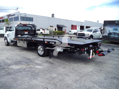 Flatbed tow truck in a dealership lot with a Chevrolet Silverado 6500HD roll-back; visible text includes stock numbers and dealership signage in background.