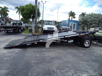Flatbed tow truck car hauler with hydraulic tilt bed in a dealership lot showing ramps and wheels.