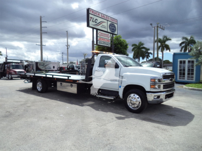 Truck with flatbed tow body in a lot; side shows no readable labels; background signage includes TLC Truck Sales Center and a blue building.