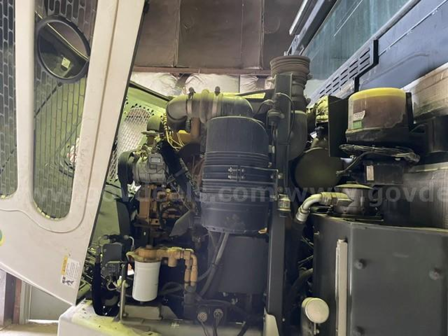 Front view of Elgin Pelican sweeper engine bay showing diesel engine, air filter, hoses, and coolant reservoir label text not fully legible.