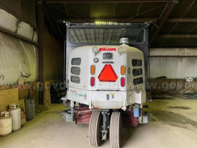 Elgin Pelican sweeper cab in a warehouse with front view showing orange reflective triangle sign and various lights. Brand 'ELGIN' on hood and warning labels near bumper.