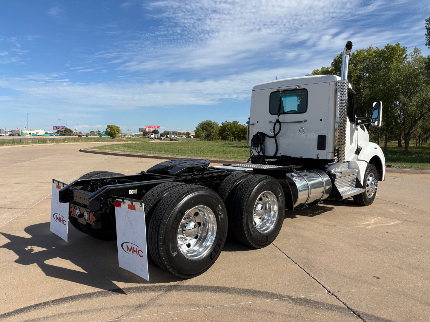Kenworth T880 tractor rear view showing dual rear axles, chrome wheels, fuel tanks, and white cab. Mudflaps with MHC logo visible.