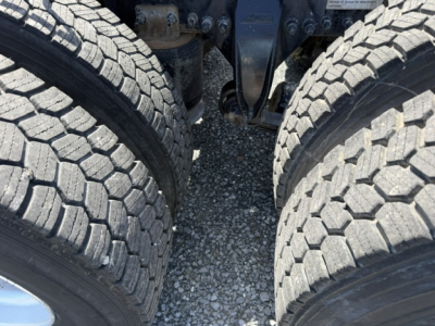 Truck chassis with dual rear axles and suspension components visible between tires; gravel surface beneath.