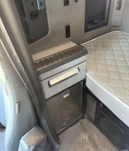 Interior of a Mack Anthem truck cab with a small grey cabinet and a two-drawer unit next to a quilted seat; control panels mounted on wall.
