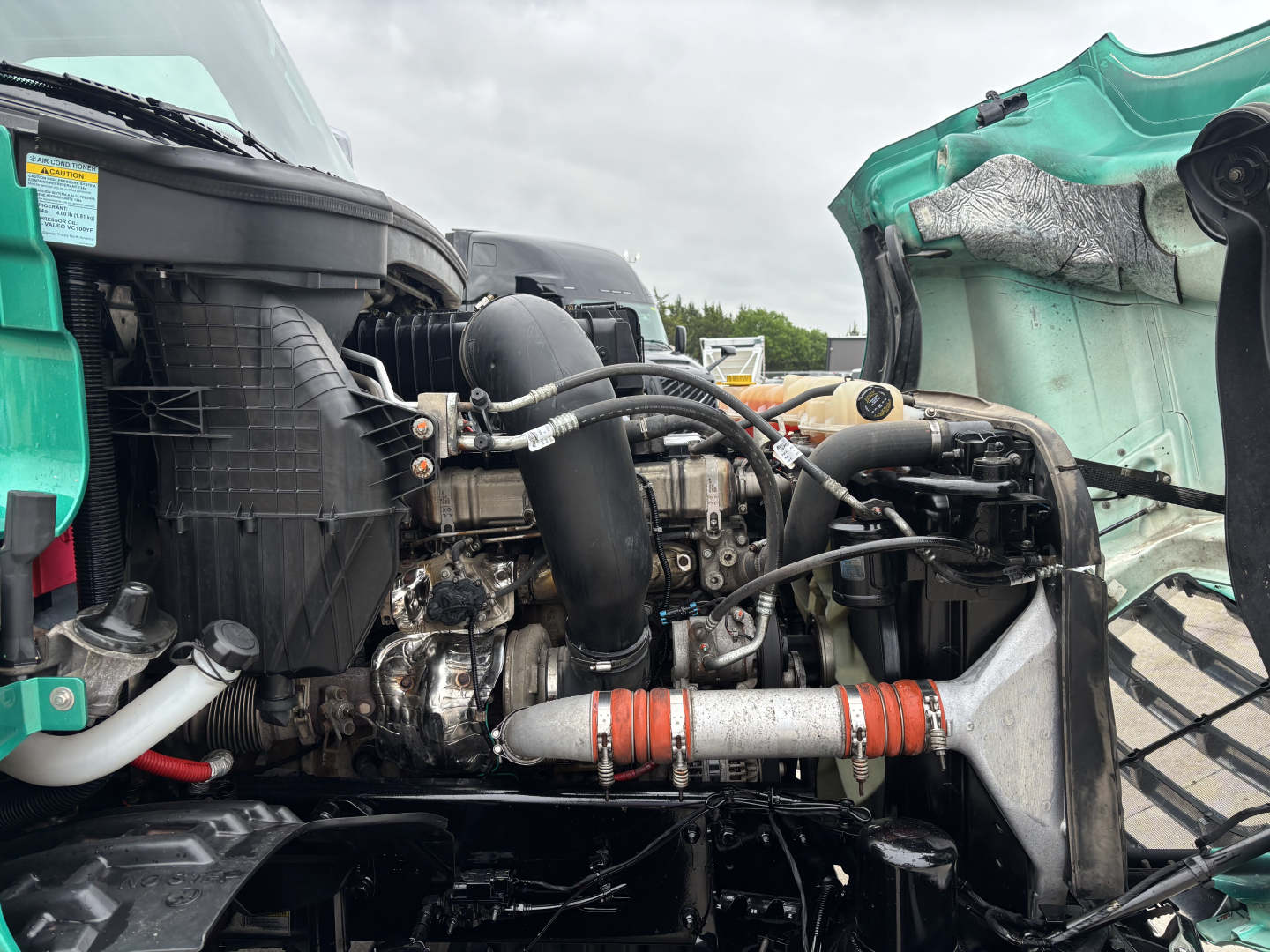 Truck engine bay of a 2023 Freightliner Cascadia 126 showing visible hoses, radiator, turbo intake, and engine components in red-orange insulated hose clamps.
